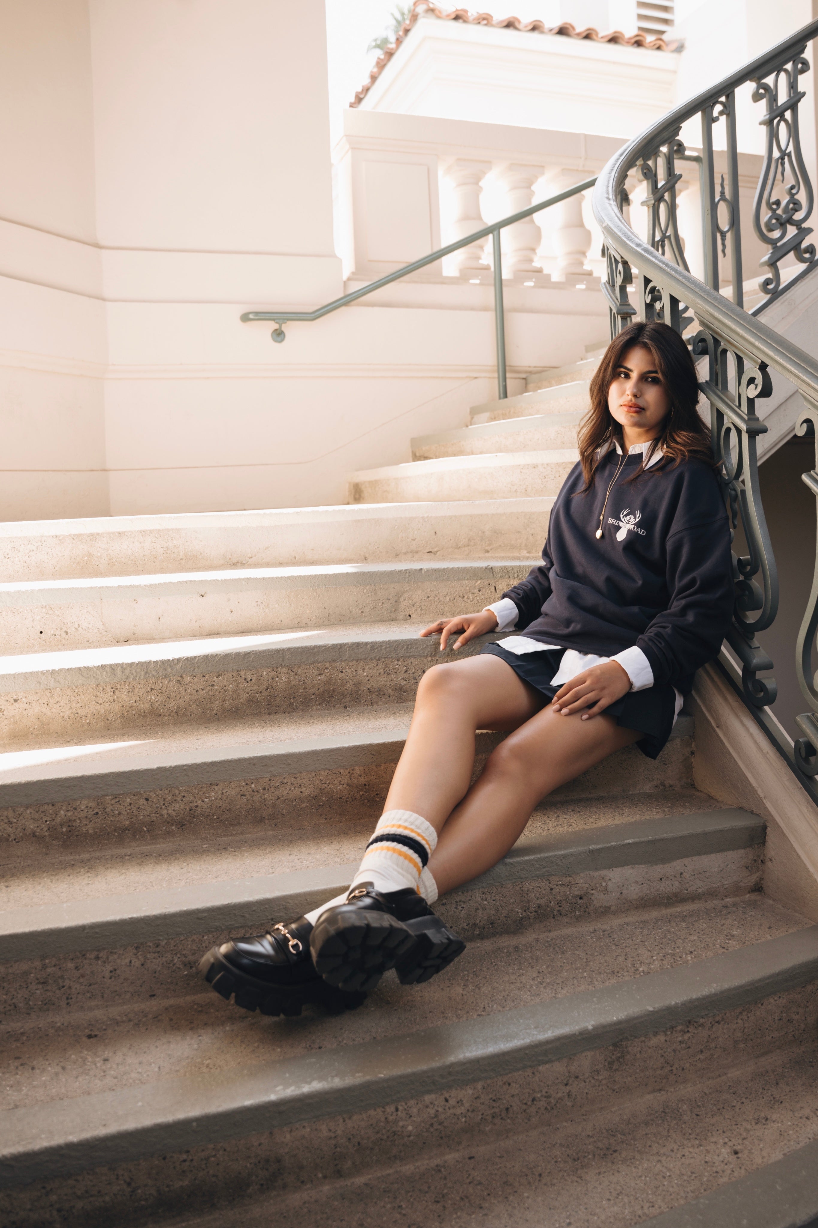 Woman posing leaning on stairs wearing grey crewneck sweater, skirt and mary jane shoes