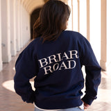 Woman facing backwards, wearing a navy blue long sleeved crewneck with Briar Road branding