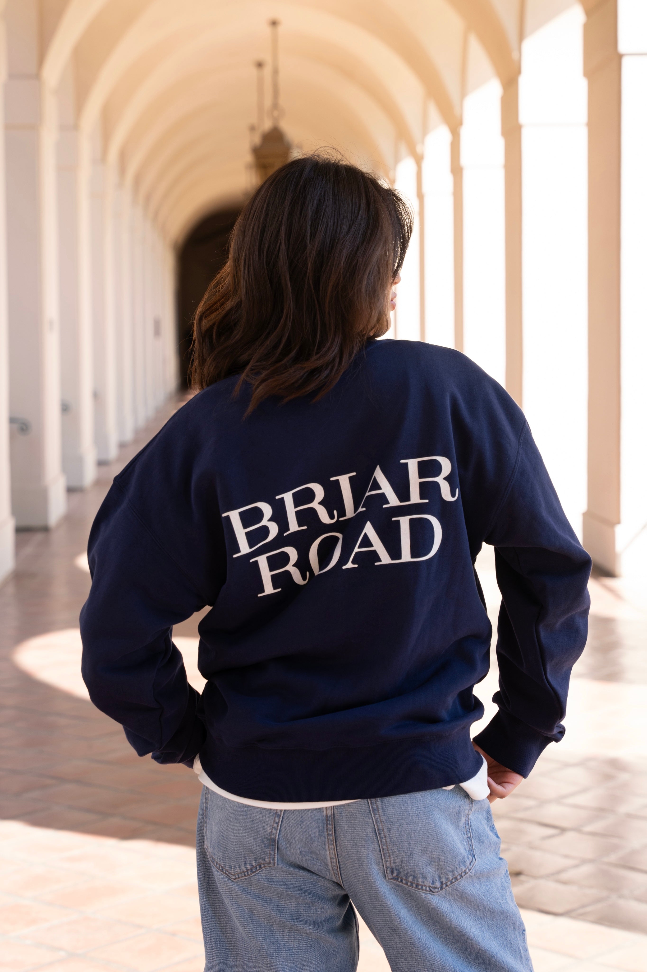 Woman facing backwards, wearing a navy blue long sleeved crewneck with Briar Road branding