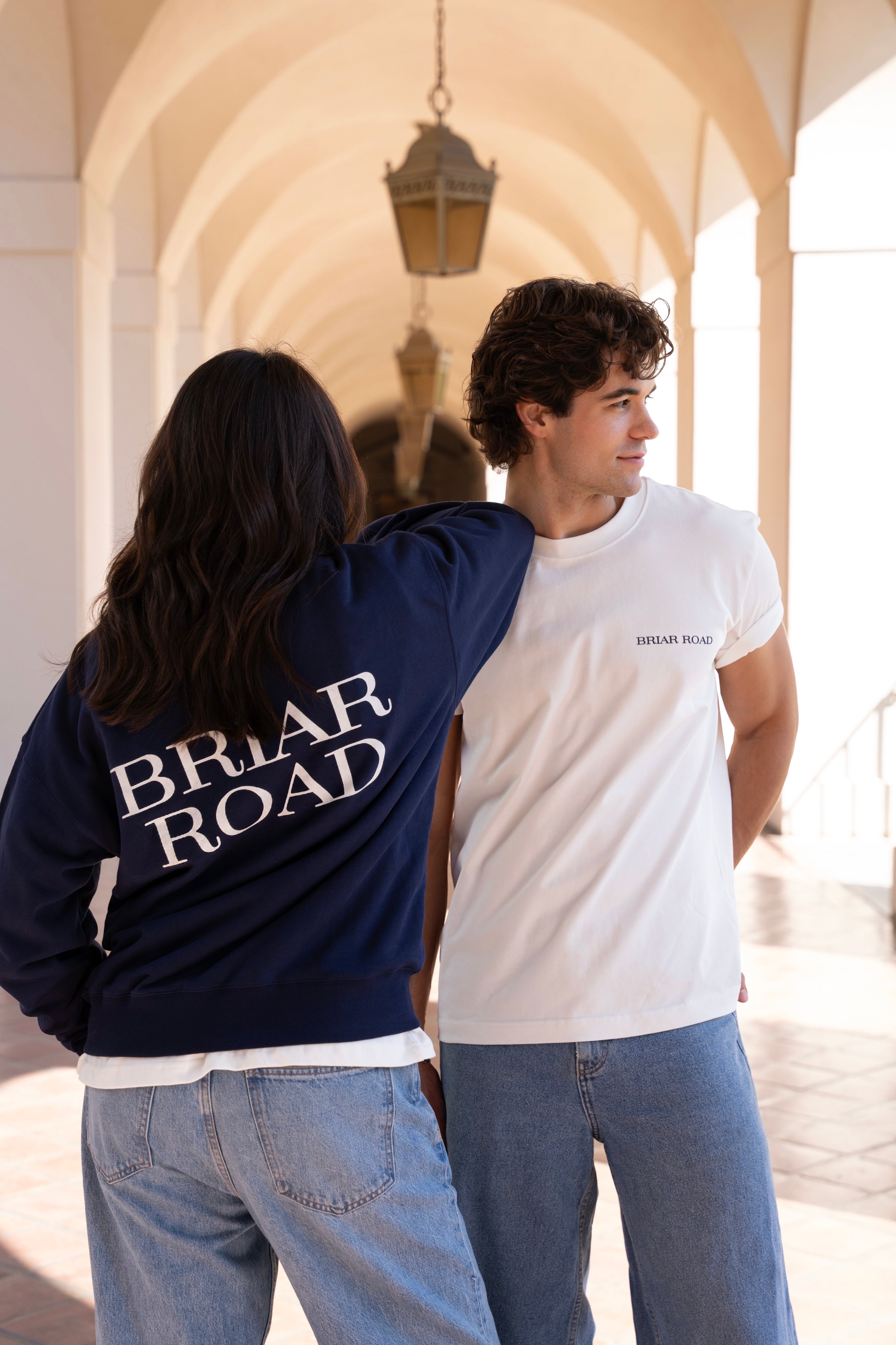 Couple posing. Woman wearing a navy blue crewneck sweater with Briar Road branding. Man wearing a white short sleeved crewneck and looking sideways.
