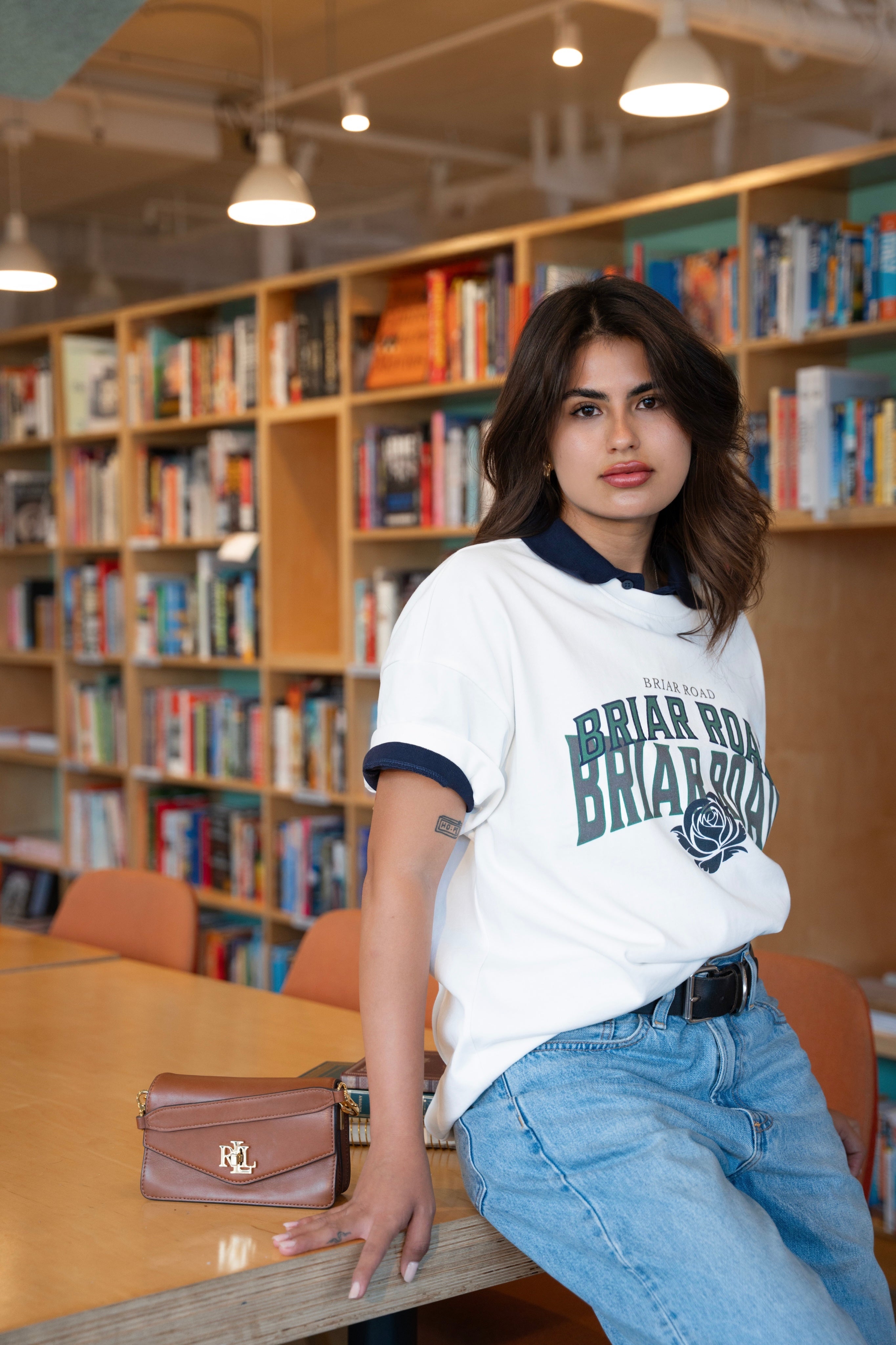 Woman leaning on a table wearing a styled white crewneck with jeans and a blue polo